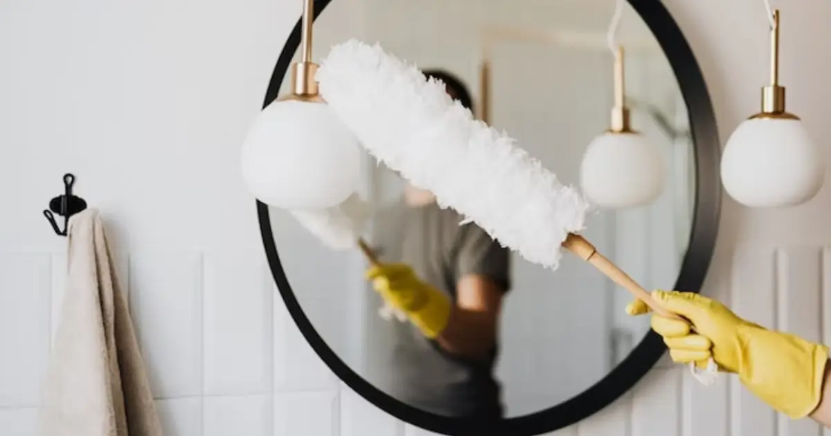 Person using a fluffy duster to clean a bathroom light fixture and mirror. Caption: A good duster is one of the essential cleaning tools every home should have.